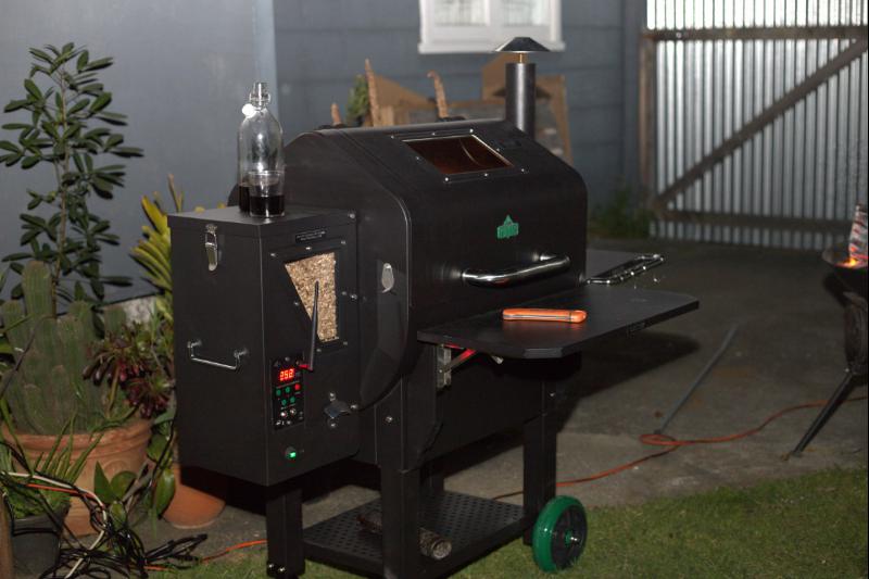 pellet grill smoker with wood pellets inside a pellet holder container as part of the smoker. It has a chimney and a bottle of  wine on top of the pellet holder on the side.