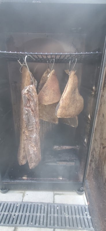 Pork meat hanging inside a smoker, with the door open and a hazy smoky view into the smoke chamber.