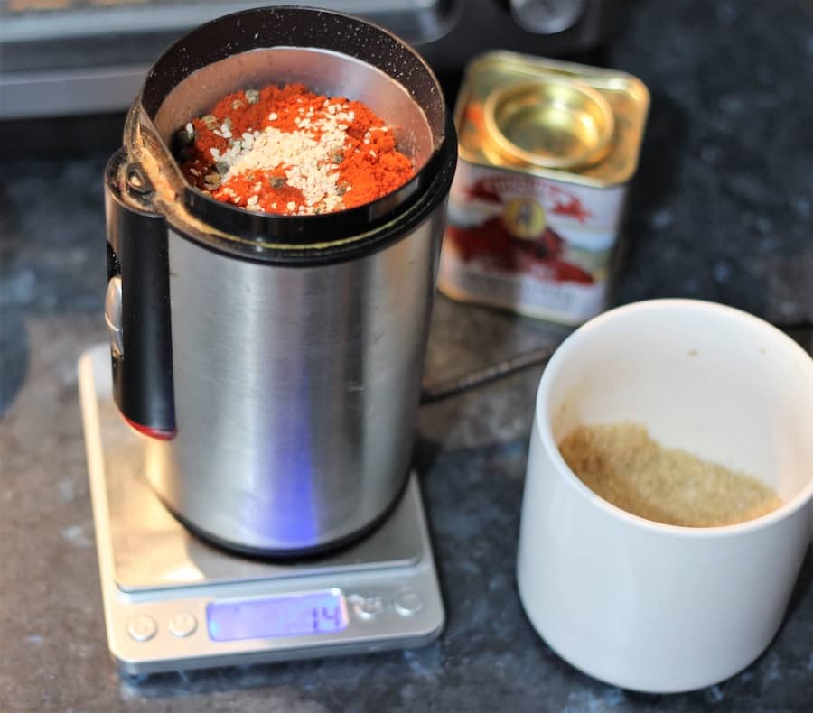 A spice grinder filled with various spices sitting on a digital kitchen scale beside a can of tomato paste and a cup of ground spice blend.