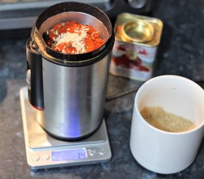 A spice grinder filled with red chili flakes and seeds on a digital kitchen scale, with a can of chili powder and a measuring cup with ground spices nearby, suggesting the preparation of a homemade spice blend as