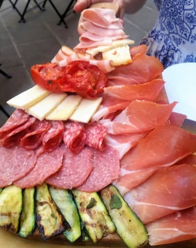 A person holding a colorful charcuterie board stacked with a variety of meats, cheese, and grilled vegetables.