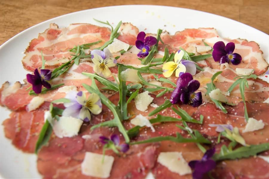 Dry Cured Salumi charcuterie dish garnished with arugula, shaved parmesan, and edible flowers on a white plate.