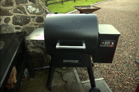 A Traeger pellet grill, one of the easiest smokers for beginners, stands outdoors on a paved surface next to a stone wall, ready for a barbecue session.