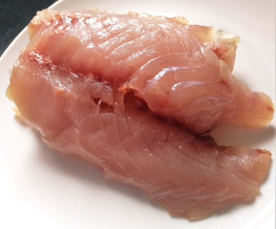 A piece of raw chicken breast on a white plate, awaiting preparation or cooking.