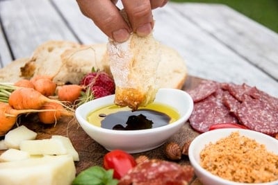 A hand dipping a piece of crusty bread into olive oil and balsamic vinegar by a rustic outdoor charcuterie board with fresh vegetables, meats, and cheese.