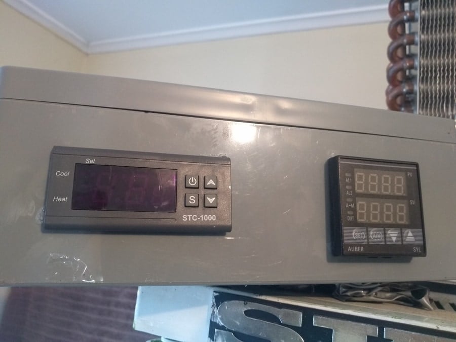 Two digital thermostats, part of the meat curing equipment, mounted on a gray surface, with one displaying a faded readout and the other showing clear numerical temperature values.