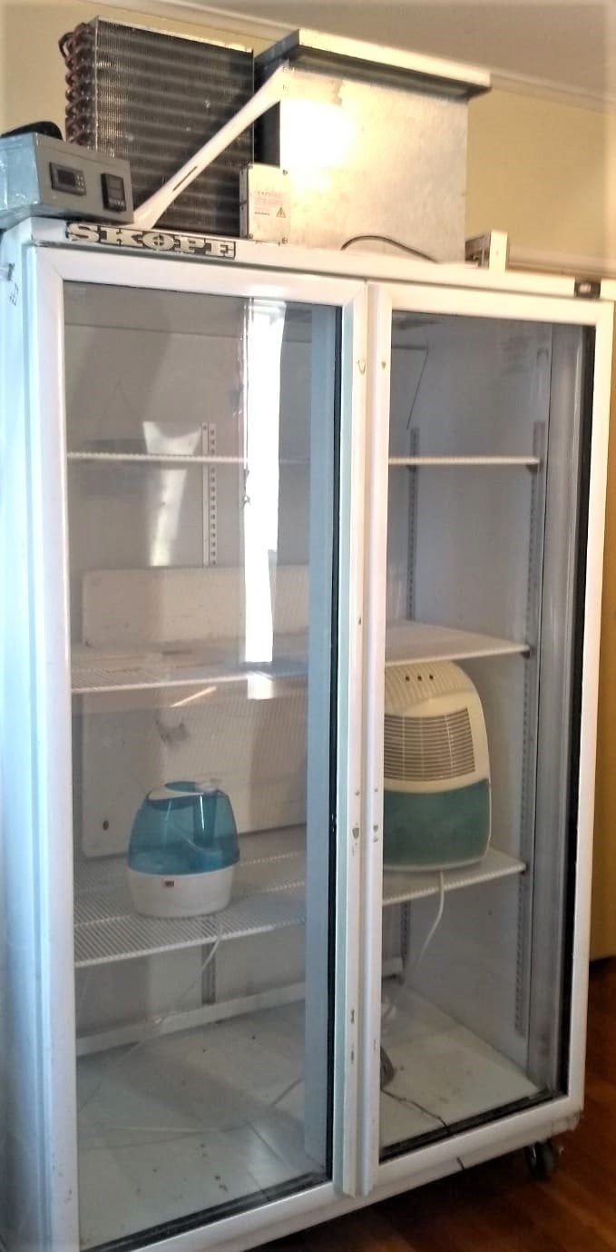 An empty commercial refrigerator with its door open, containing only a few small items including a portable air cooler, a water jug, and a drying cabinet on its shelves.