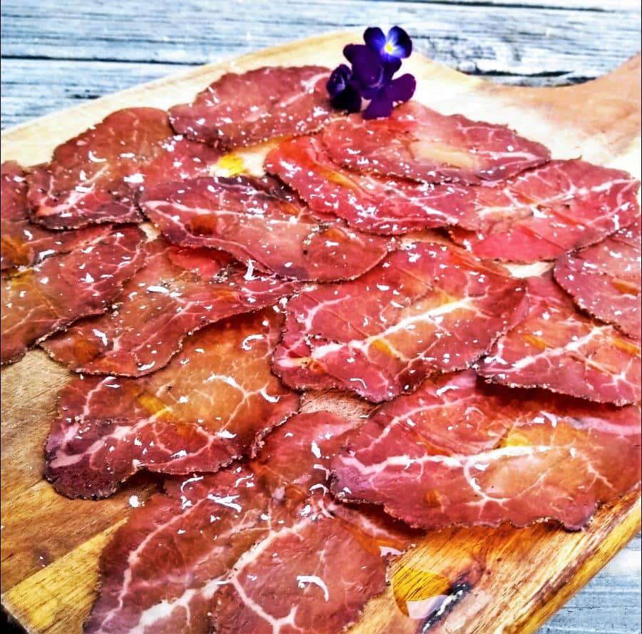 Thinly sliced cured meats garnished with a violet flower, elegantly presented on a wooden cutting board.