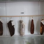 Various cured meats hanging in a normal fridge with tags, possibly indicating type or date of curing.