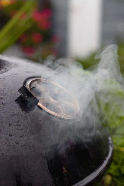 A smoking grill outdoors on a rainy day, with steam rising from the lid indicating something delicious cooking inside.