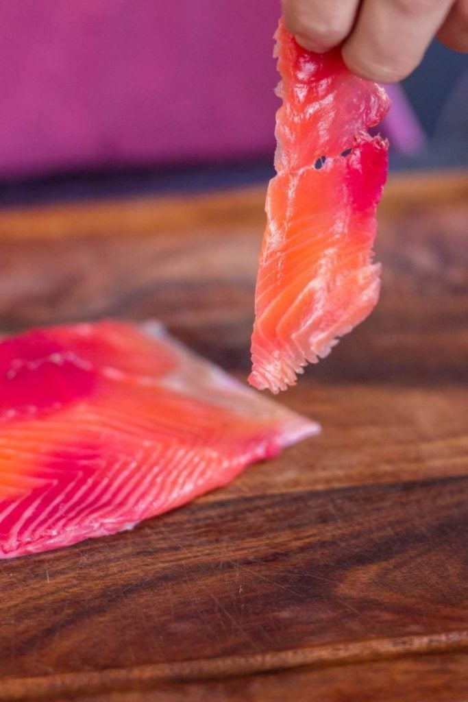 A close-up of a person's fingers holding a translucent, thin slice of smoked salmon over a wooden surface.