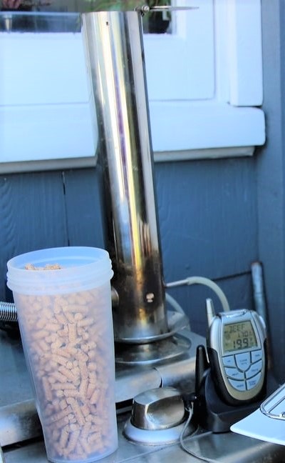 A Beginners Guide to a pellet smoker setup with a container of wooden pellets, a digital temperature control, and smoking meat equipment against a house exterior.