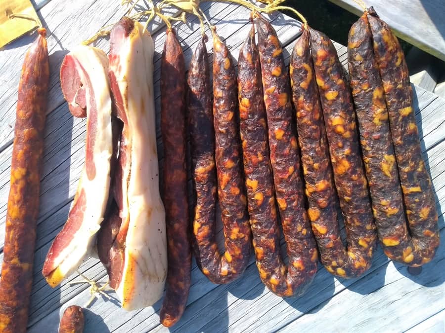 A selection of cured meats, including sausages and bacon, hangs on a wooden surface to dry in the sunlight, exploring food preservation techniques.