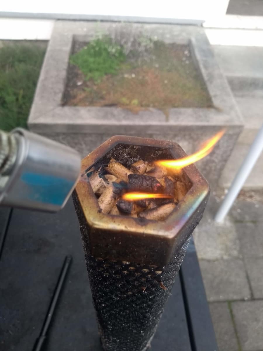 Igniting a portable biomass stove, with a visible flame heating the pellet tube inside as wisps of smoke rise.