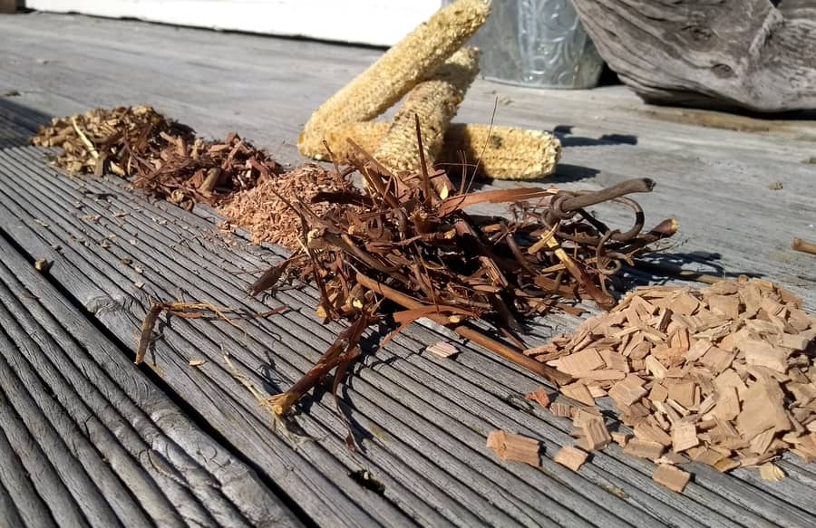 Various natural textures: corn cob, dried leaves, twigs, and smoking wood shavings scattered on a wooden deck.