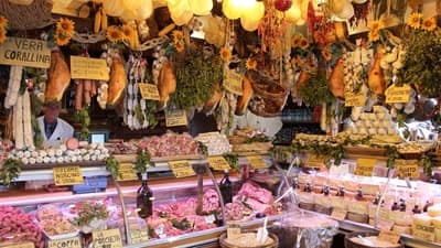 A bustling deli displaying an abundance of fresh meats, cheeses, and other delicatessen products, with a vendor attending to the goods amidst warm lighting and vibrant decorations.