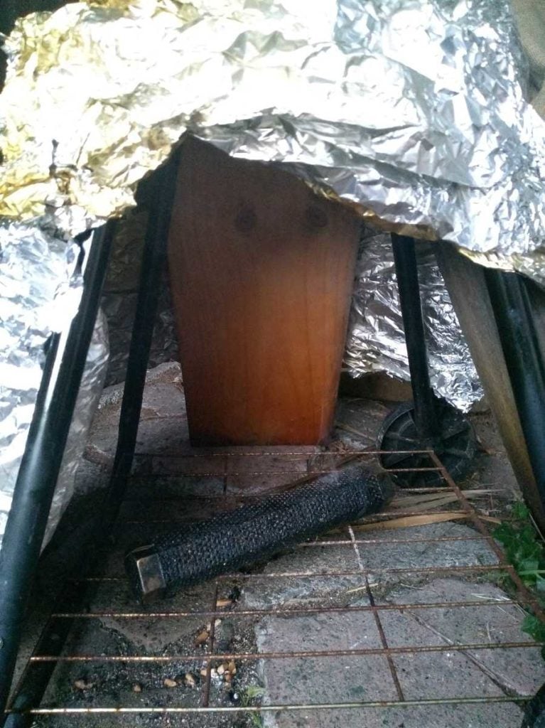 A makeshift wooden smoker covered with aluminum foil cooking food outdoors, with a visible log of wood generating smoke for flavoring.