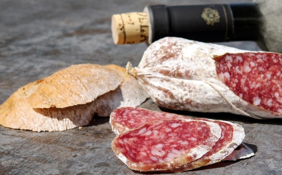 A rustic gourmet snack with sliced cured meat, crusty bread, and a bottle of wine with a cork.