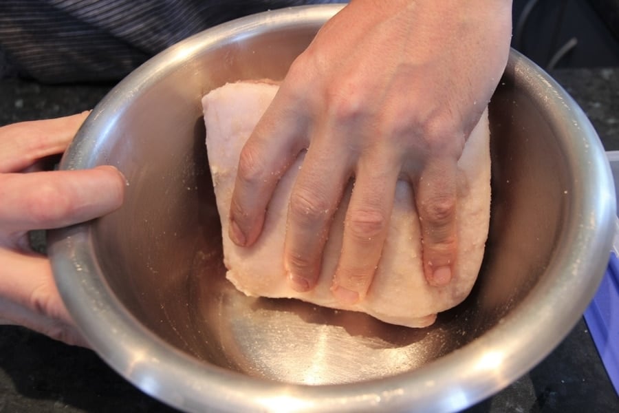 A person's hands pressing cured meat in a stainless steel bowl on a kitchen counter.