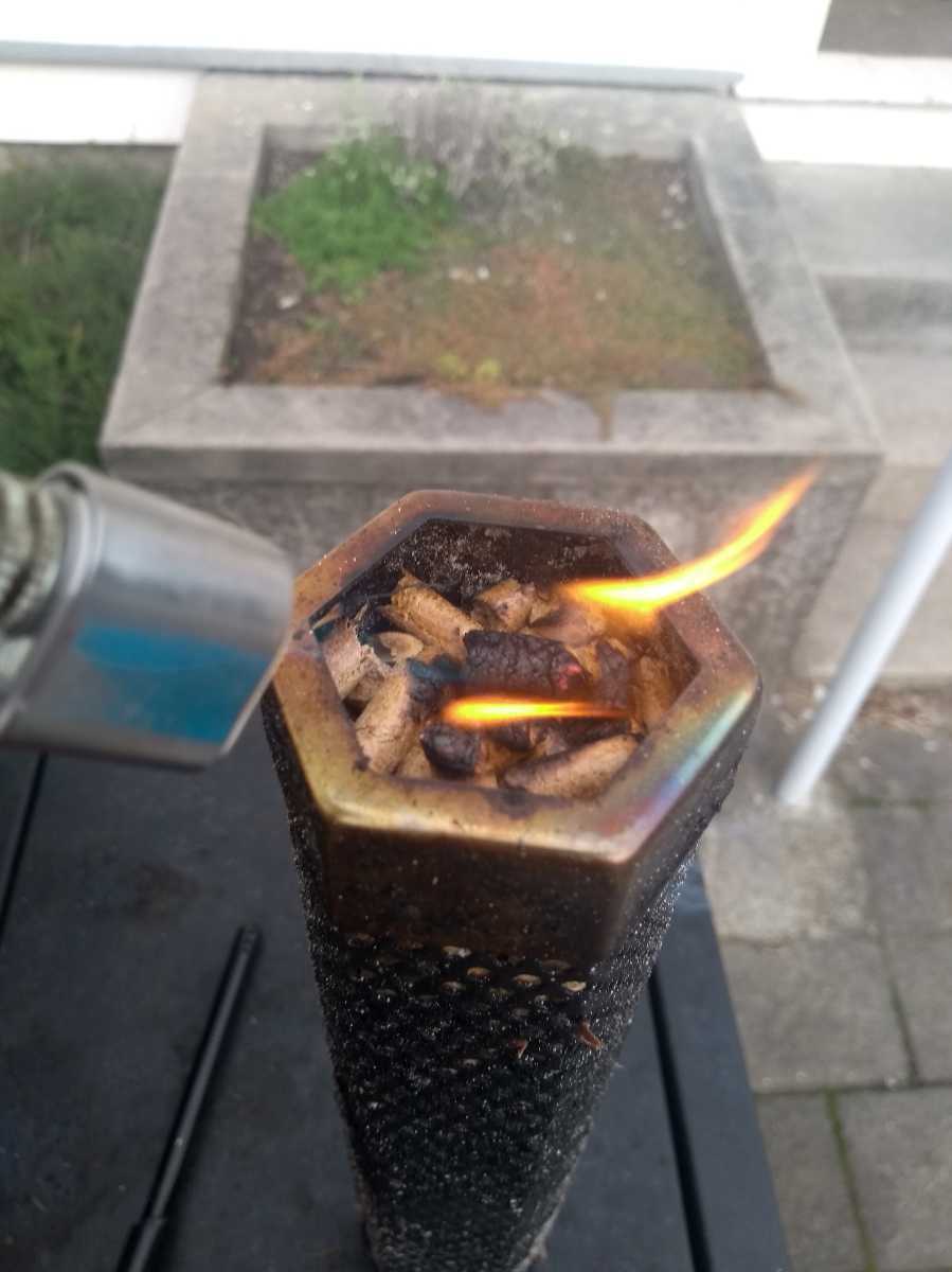 A close-up of a handheld butane torch flame heating a metal device filled with brown contents, possibly a smoking accessory, against an outdoor background.