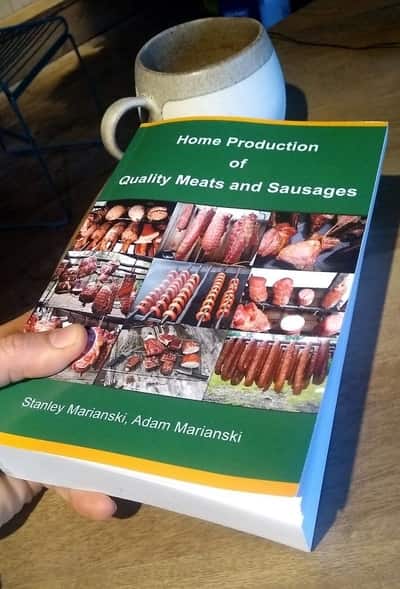 A hand holding a book titled "Home Production of Quality Meats and Sausages" by Stanley Marianski and Adam Marianski, focusing on meat curing and dry curing, with a cup