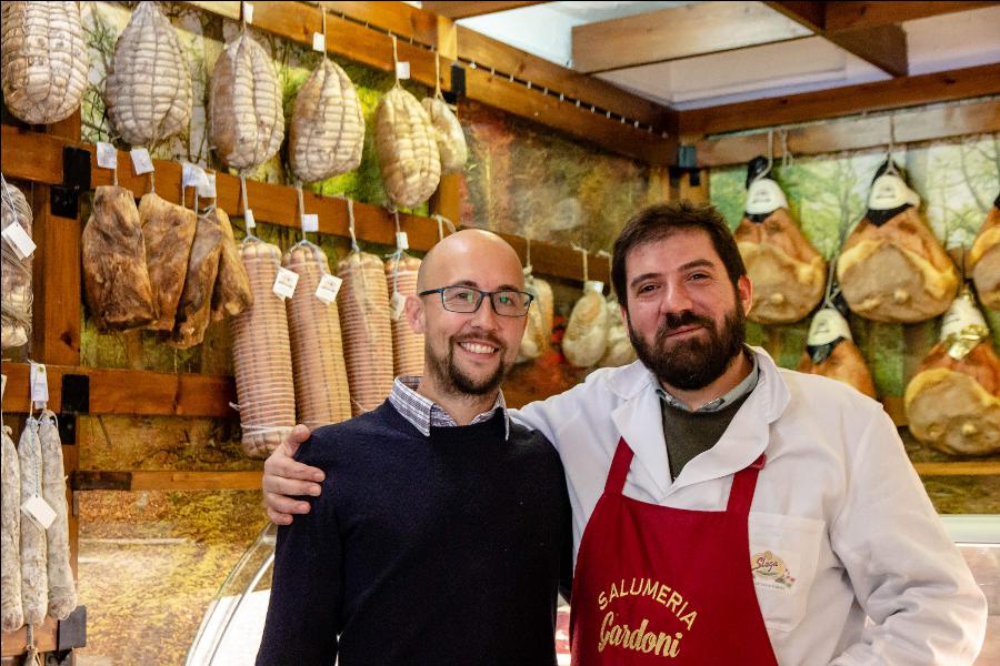 Two smiling men, one in casual attire and the other wearing an apron, stand proudly in front of a display of hanging cured meats in a traditional delicatessen.