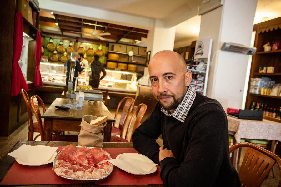 A man seated at a table in a cozy deli, preparing to enjoy a plate of sliced cured meats.