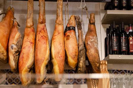 Cured hams hanging in a traditional storage, with a selection of wine bottles in the background, suggesting a well-stocked pantry or a specialty food shop.