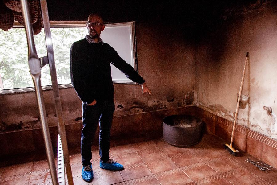 A man standing inside a room with worn-out walls pointing towards a large pot, with a broom nearby and natural light coming in through a window.