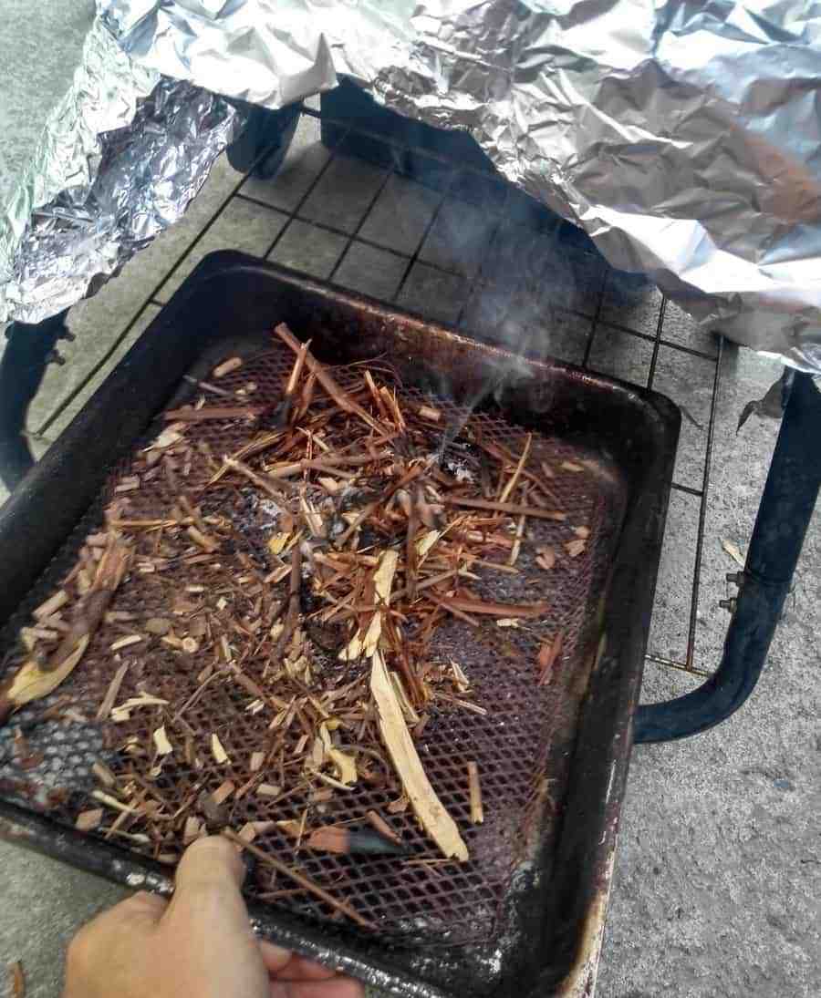 A person is lighting a barbecue grill covered with wood chips for smoking foods, with a protective aluminum foil tent set up over it.