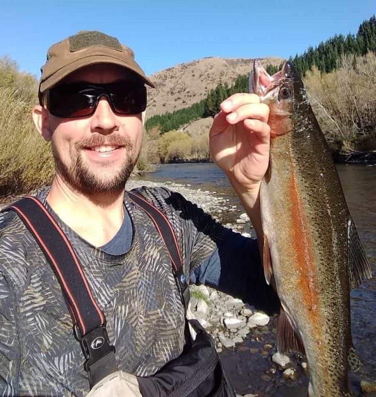 A smiling fisherman wearing a cap and sunglasses proudly holding up his trout catch of the day by the river.