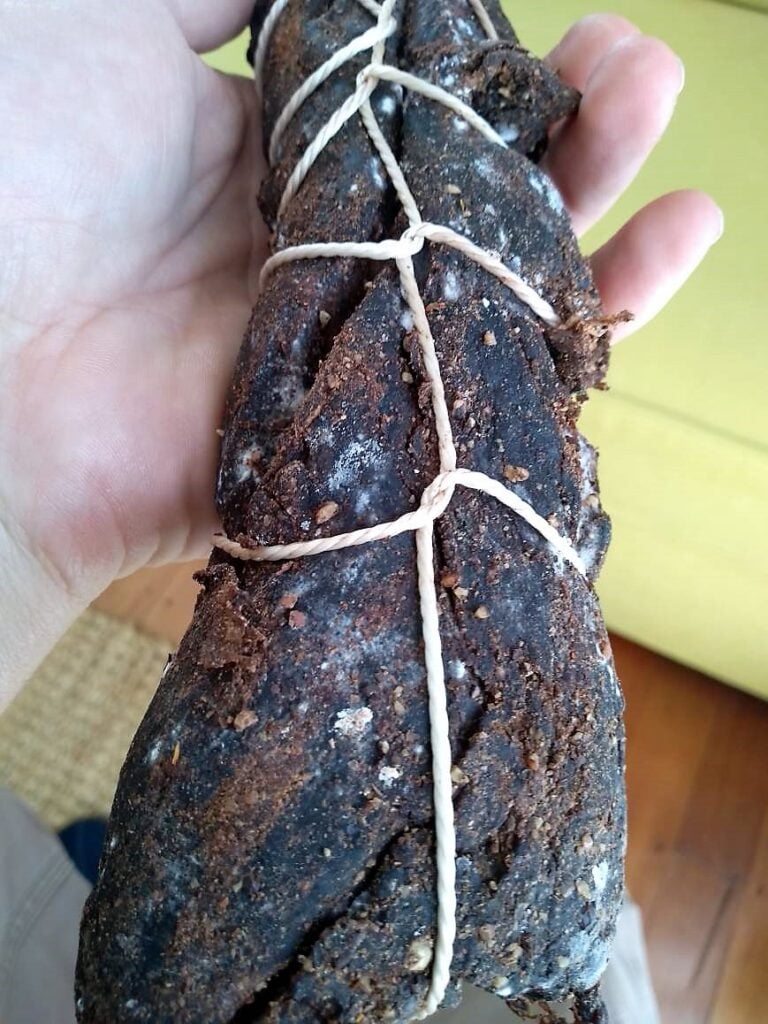 A close-up of a hand holding a large, rustic-looking sausage tied with string, possibly homemade or artisanal.