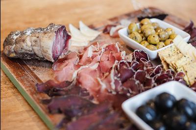 A selection of charcuterie and olives on a wooden board, featuring thinly sliced cured meats, a piece of rustic bread, and bowls of green and black olives.