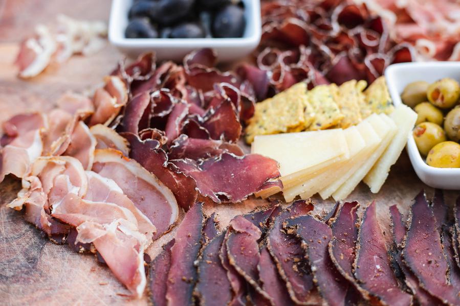 Salt Dried Cured Meat in a wooden board. Thin Slices of Venison (Pastirma) and Pork Loin. With some charcuterie board condiments - crackers, cheese and olives