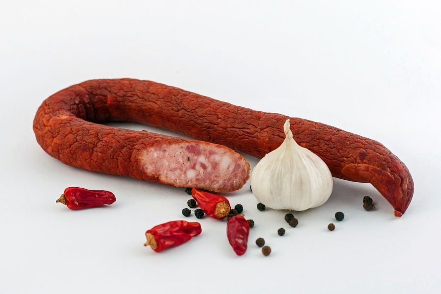A smoked salami with a slice cut out, alongside a whole garlic bulb, some dried red chili peppers, and scattered black peppercorns on a white background.