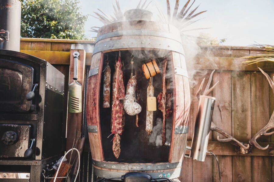 Traditional meat cold smoking process in a homemade barrel smoker with various cuts of meat hanging inside and smoke rising from the smoker, detailed in our comprehensive smoking eBook.