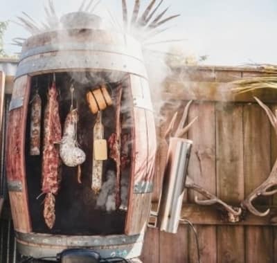 A homemade meat smoker, now a piece of essential equipment, crafted from an old beer keg and surrounded by rustic wooden walls, emanates smoke as it performs cold smoking on a selection of meats and