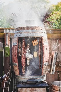 Rustic outdoor smoker in action, with aromatic smoke billowing out as meat and cheese hang inside, infusing with rich flavors thanks to the integrated cold smoke generator.