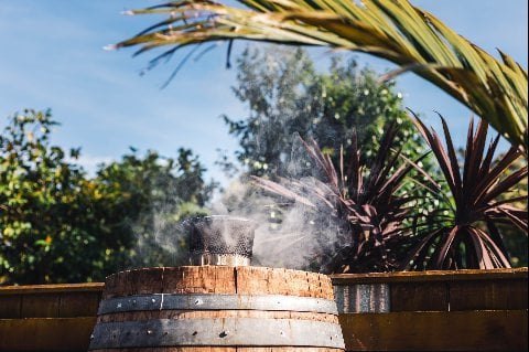 A rustic wooden barrel basks in the outdoor sunshine as wisps of smoke rise gently from a smoldering object on top, surrounded by lush palm foliage under a clear blue sky.