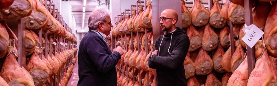 Two people discussing amidst rows of hanging cured meats in a storage facility or aging room.