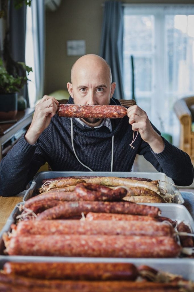 A man playfully pretending to bite into a long sausage link of cured meat with trays of freshly made sausages in front of him.