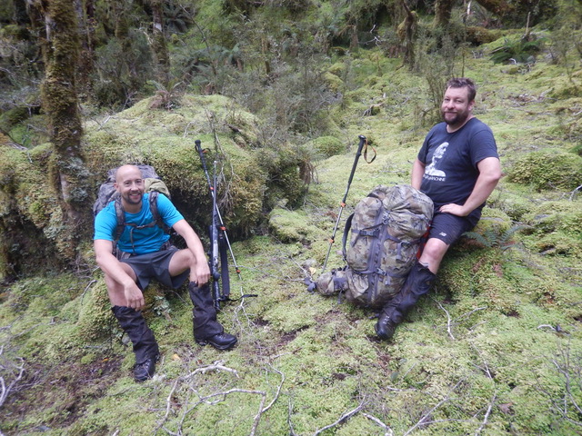 Two hikers, including Tom Mueller, taking a break in a lush green forest, surrounded by moss-covered ground with trekking poles and a heavy backpack.