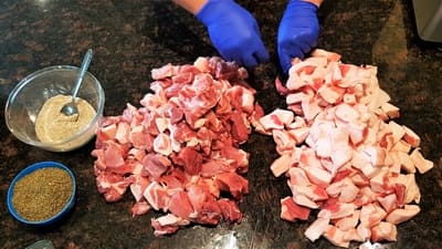 Hands in blue gloves preparing a large quantity of diced raw meat and pork fat on a kitchen countertop, with seasoning and a bowl of sauce nearby.