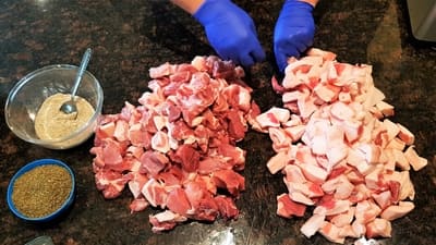 Hands in blue gloves preparing cubed pork fat on a kitchen countertop, with a bowl of seasoning on the side.