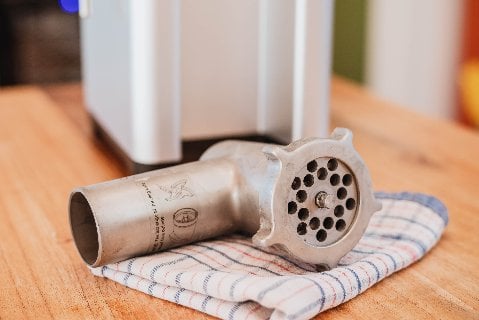 A shiny metal meat grinder attachment resting on a striped kitchen towel, with a blurred background featuring kitchen counters and appliances.