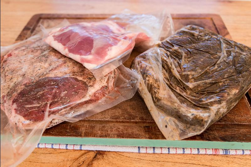 Vacuum-sealed steak and cured bacon resting on a wooden cutting board, likely marinating or being prepared for sous vide cooking.