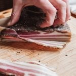 Slicing through layers of flavor: a hand carefully cuts thin slices of pancetta on a wooden board.