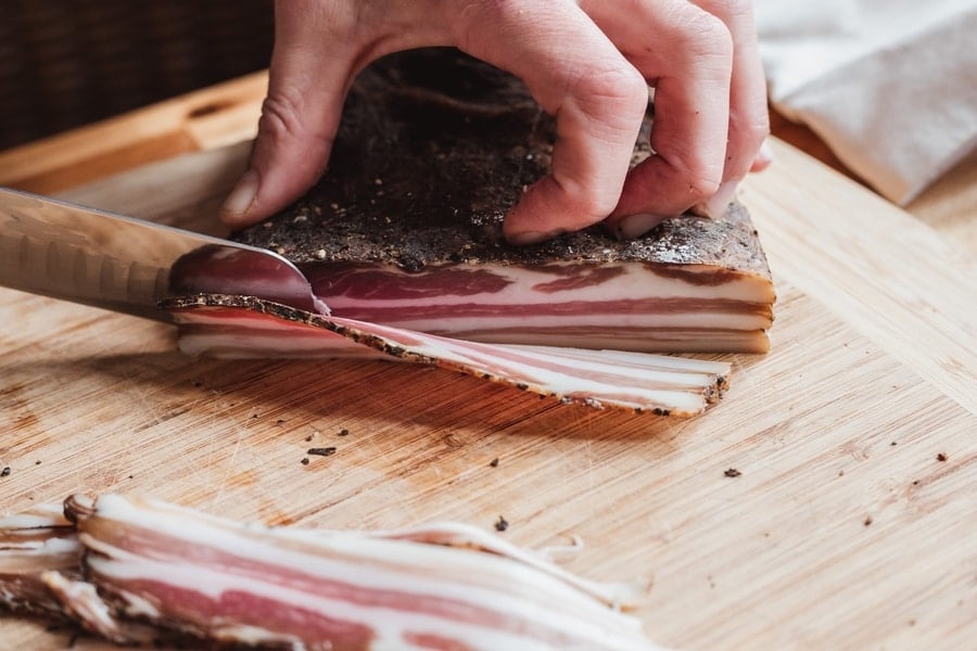 Slicing through layers of flavor: a hand carefully cuts thin slices of pancetta on a wooden board.