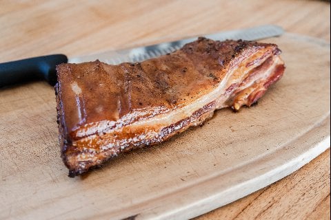 Grilled cured pork belly on a wooden cutting board with a knife nearby.