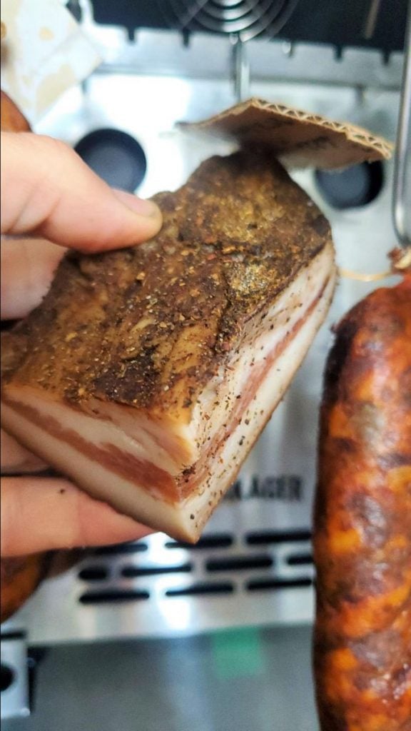 A close-up of a hand holding a slab of seasoned bacon ready to be cooked in the SteakAger Pro 40, with other meats prepped for charcuterie in the background, likely in